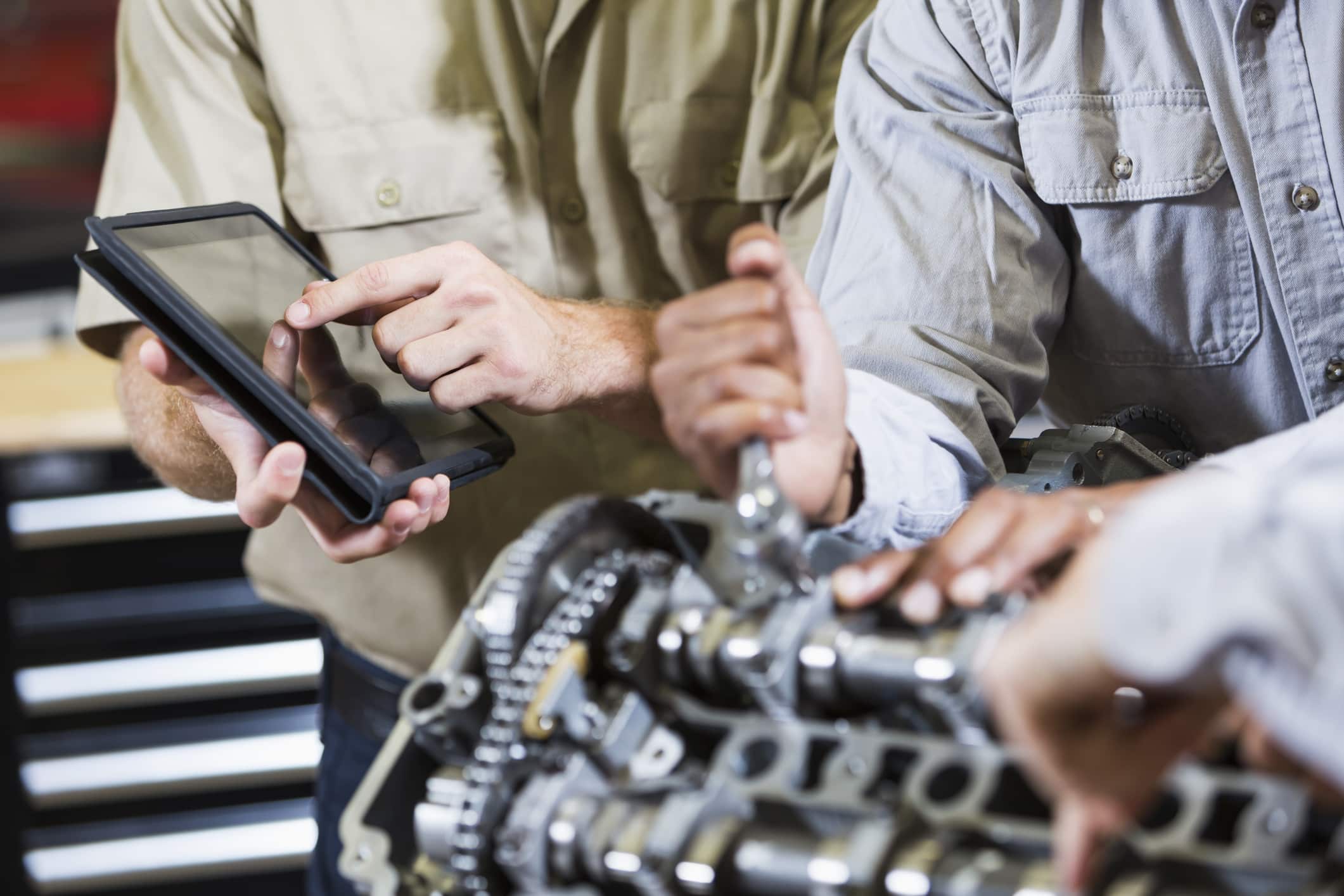 Cropped view of a group of mechanics working on a car engine. The men are unrecognizable, with arms and midsections showing. The man on the left is using a digital tablet.